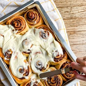 Spreading cream cheese frosting on a baking pan of overnight cinnamon rolls