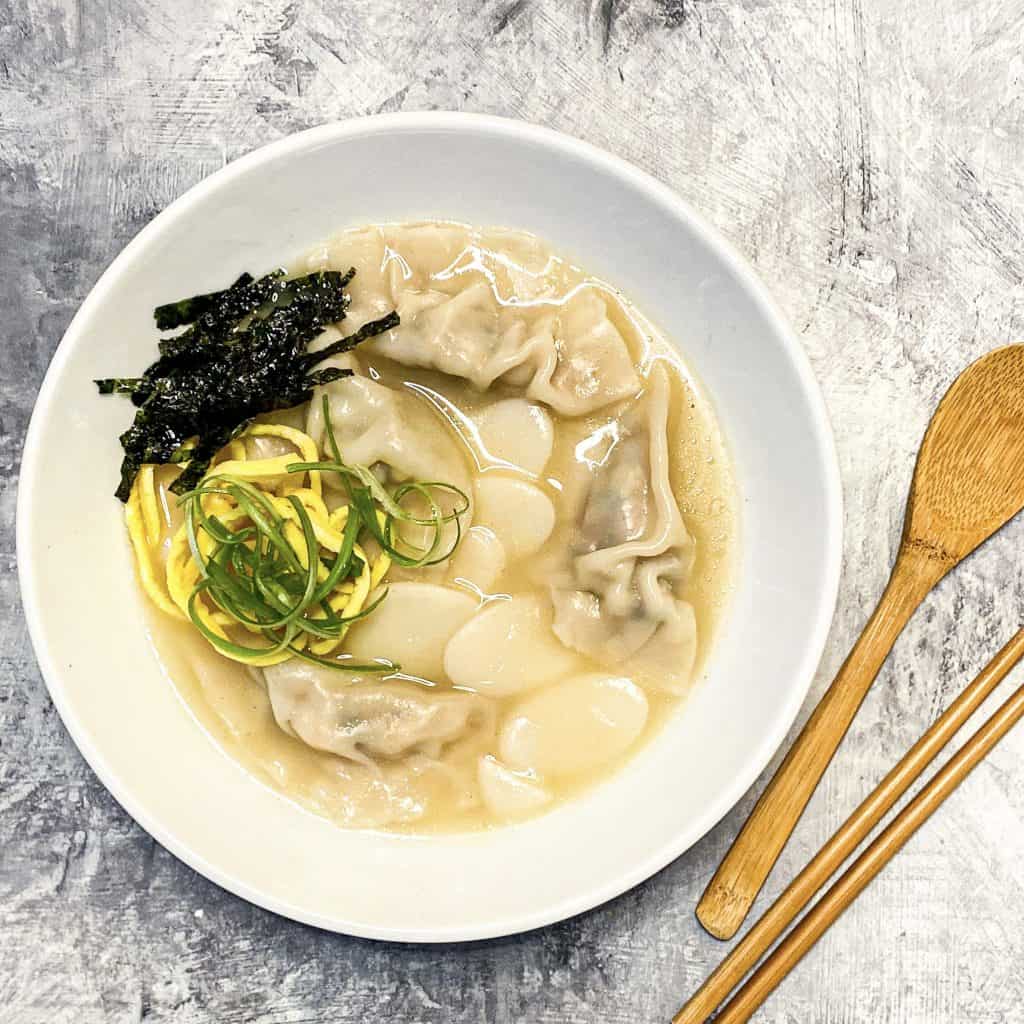 Tteok Mandu Guk (Korean Rice Cake and Dumpling Soup) in a white bowl with chopsticks and wooden spoon)