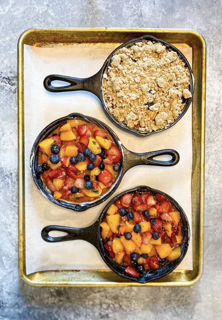 Three mini cast iron pans filled with mixed fruit, one with an oat crisp spread on top.