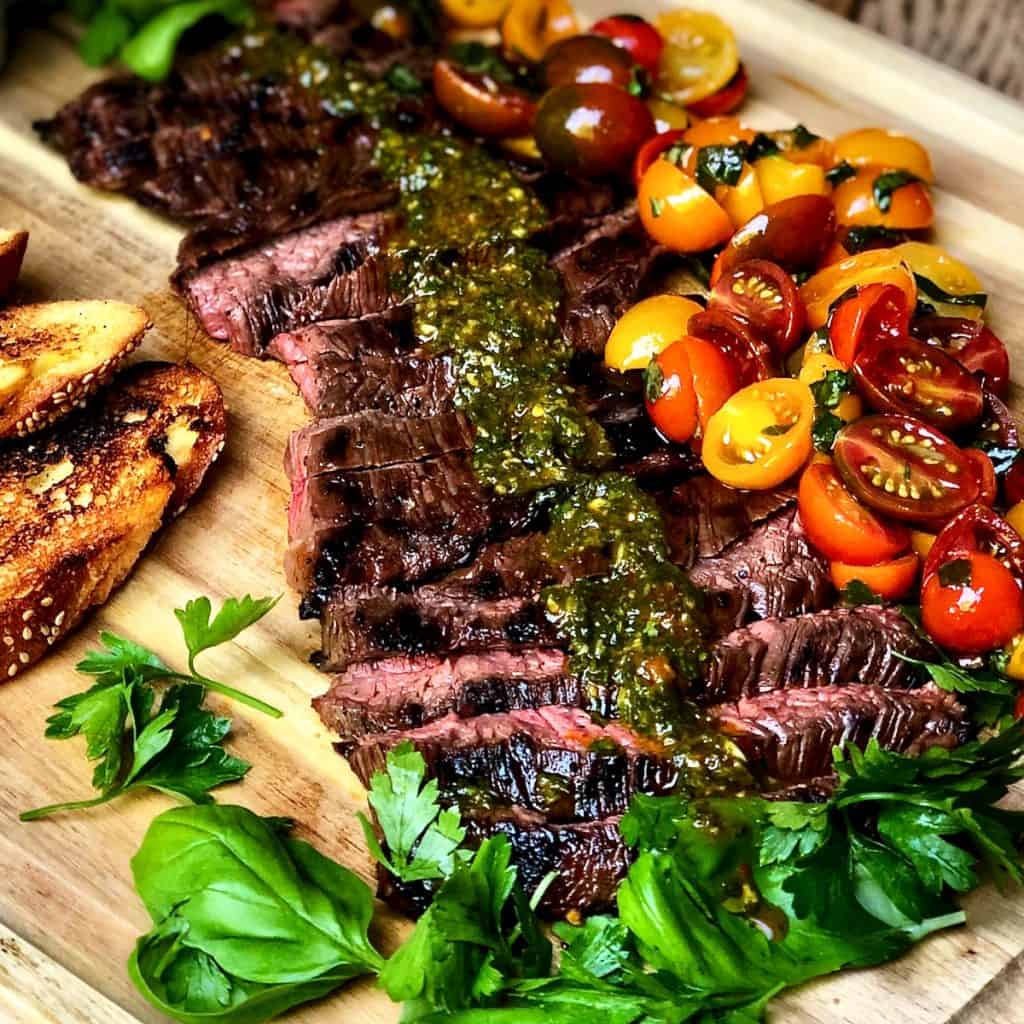 A wooden cutting board with toasted bread and Skirt Steak with Italian Chimichurri and Tomato Salad