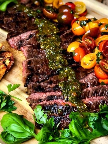 A wooden cutting board with toasted bread and Skirt Steak with Italian Chimichurri and Tomato Salad