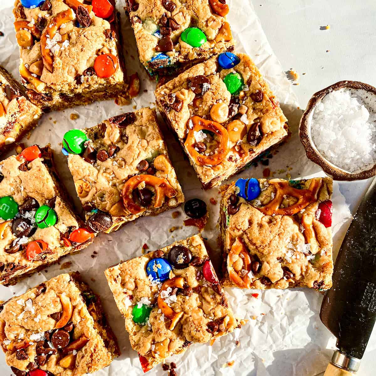 Brown Butter Blondies with M&Ms, pretzels, chocolate and peanut butter chips with a knife and flaky salt beside them.