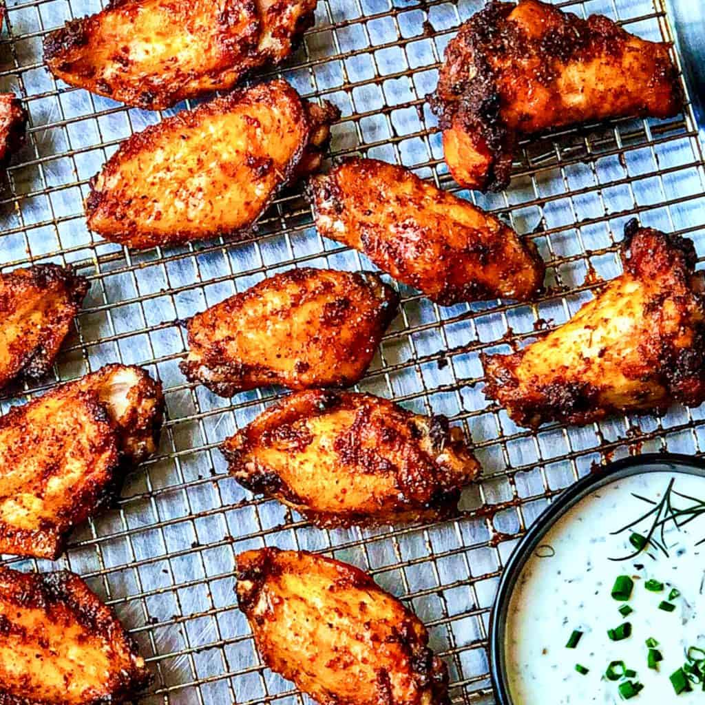 A wire rack, set in a sheet tray, covered in dry rub wings and a bowl of ranch dressing.