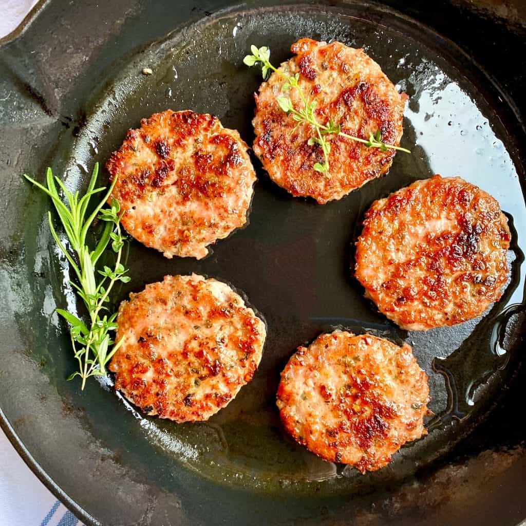 Ground Breakfast Sausage patties in a cast iron skillet with fresh herbs.