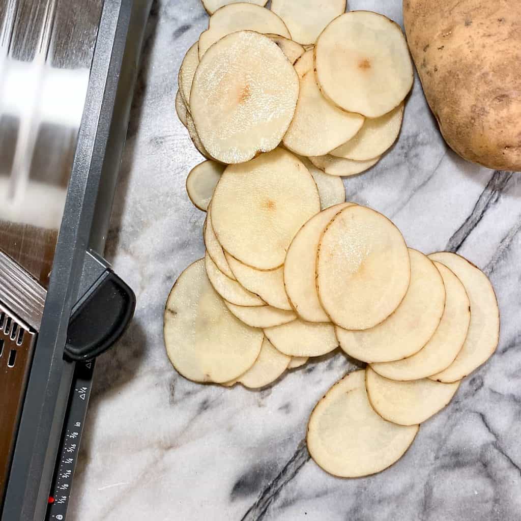 A mandolin, russet potato, and a pile of very thinly sliced raw potato on a marble board.