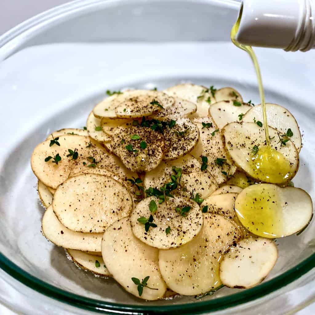 A glass bowl filled with very thinly sliced raw potatoes, seasoned with olive oil, salt, pepper, and fresh thyme.