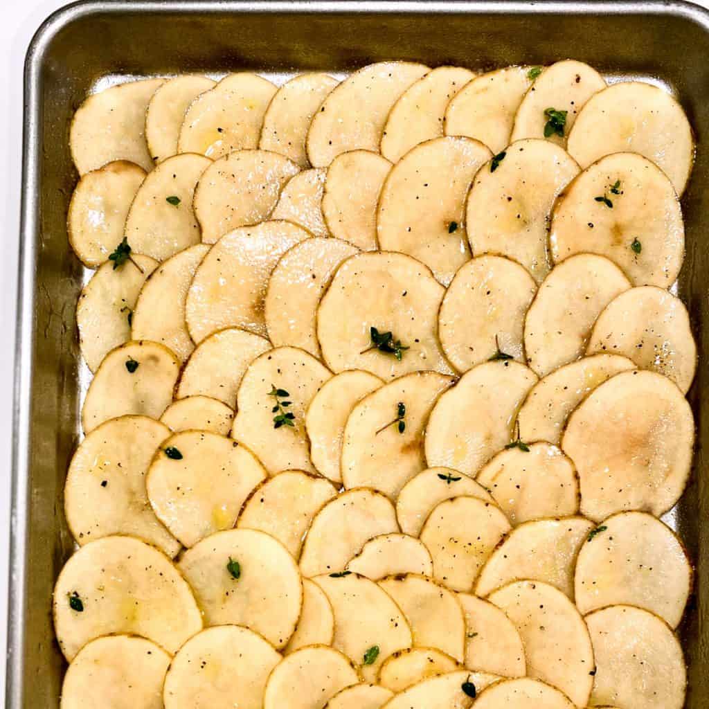 Very thinly sliced raw potatoes on a sheet pan, seasoned with olive oil and fresh thyme.