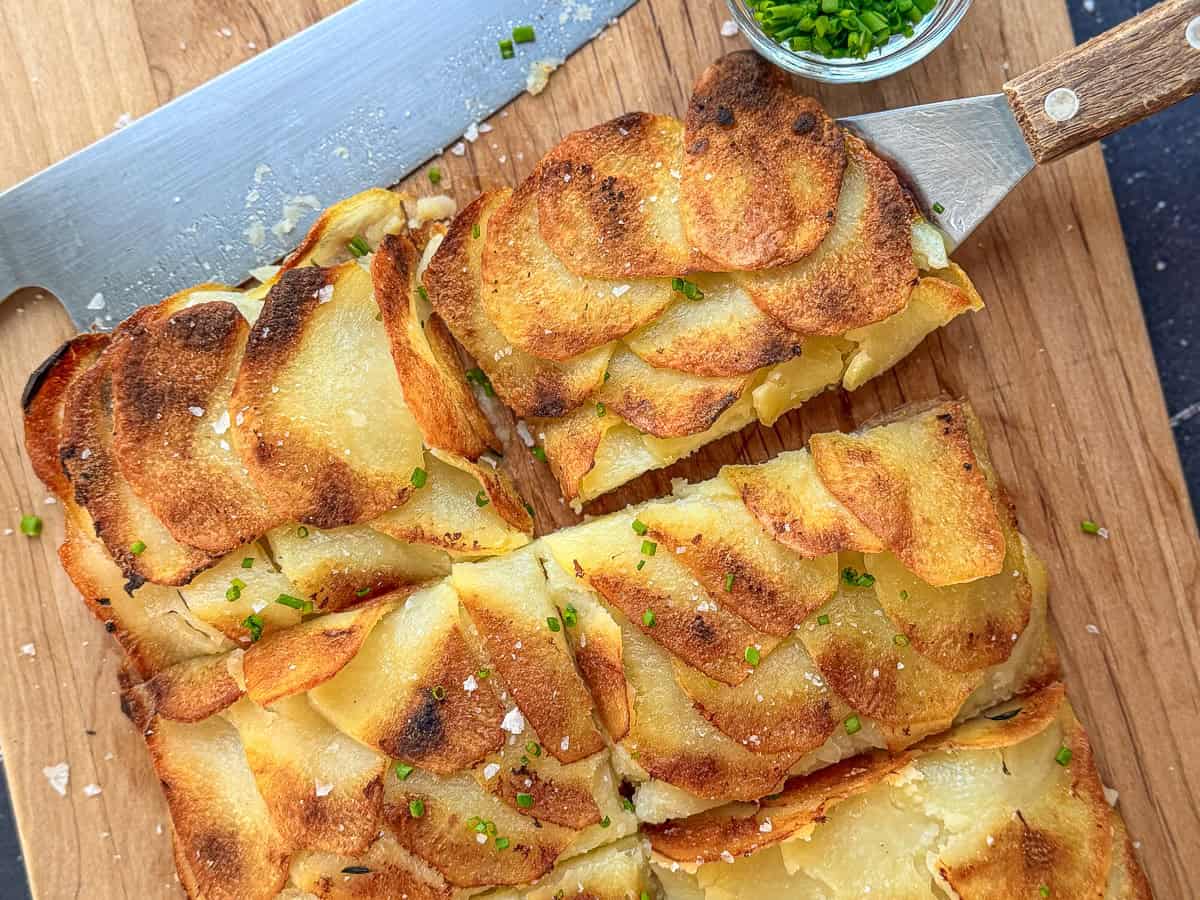 Sliced pommes anna on cutting board with chives and a knife.