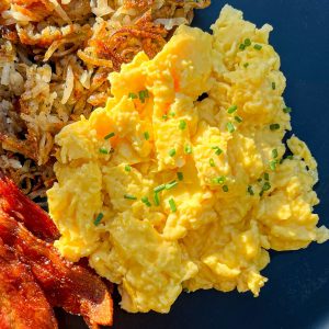 Scrambled eggs, sprinkled with chives, on a black plate with bacon and hash browns.