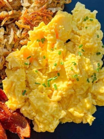 Scrambled eggs, sprinkled with chives, on a black plate with bacon and hash browns.