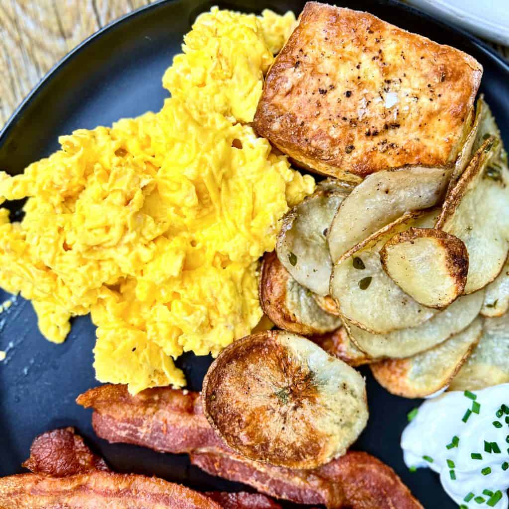Black plate containing scrambled eggs, biscuits, potatoes, and bacon.