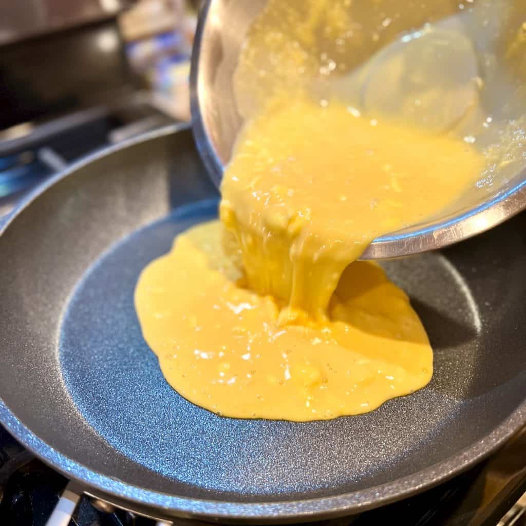 Scrambled egg mixture being poured out of a silver bowl into a nonstick pan.