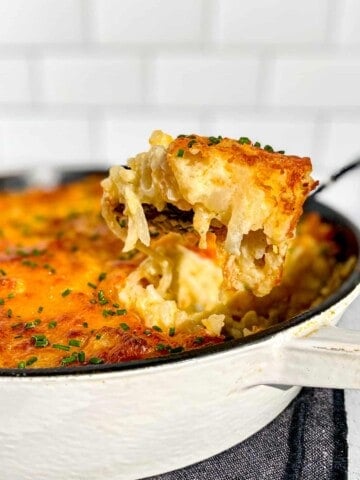 Taking a spoonful of cheesy hashbrown casserole out of a white cast iron pan.
