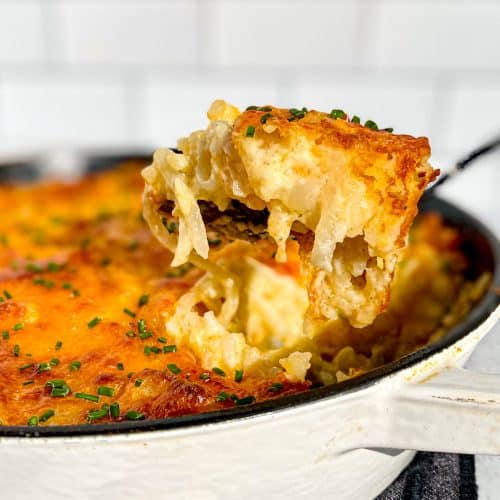 Taking a spoonful of cheesy hashbrown casserole out of a white cast iron pan.