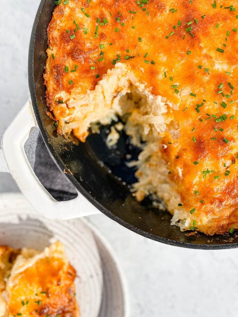 A white cast iron skillet, and a small white plate, containing cheesy hash brown casserole.