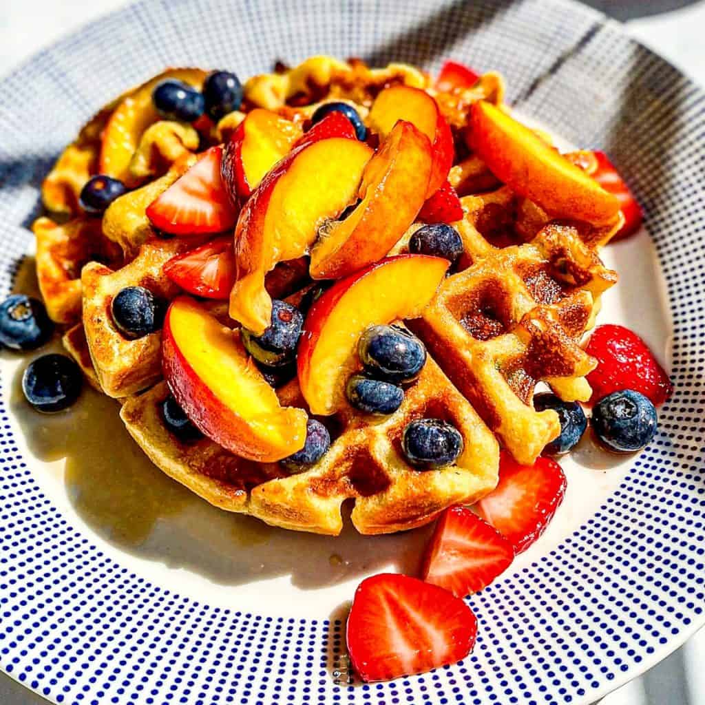 A cornmeal waffle on a blue dotted plate, topped with peaches, strawberries, and blueberries.