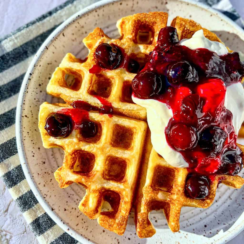 Buttermilk waffles on a white plate, topped with whipped cream and blueberry sauce.