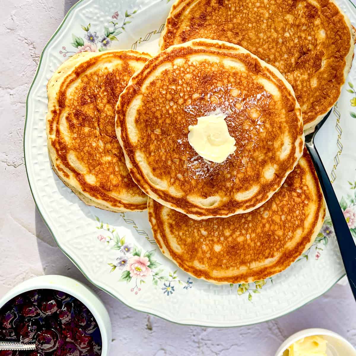 3 pancakes on a flowered plate with butter and syrup and a black serving spatula.