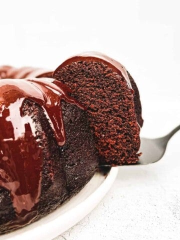 A chocolate bundt cake ona large plate with a cake cutter holding a slice of cake.