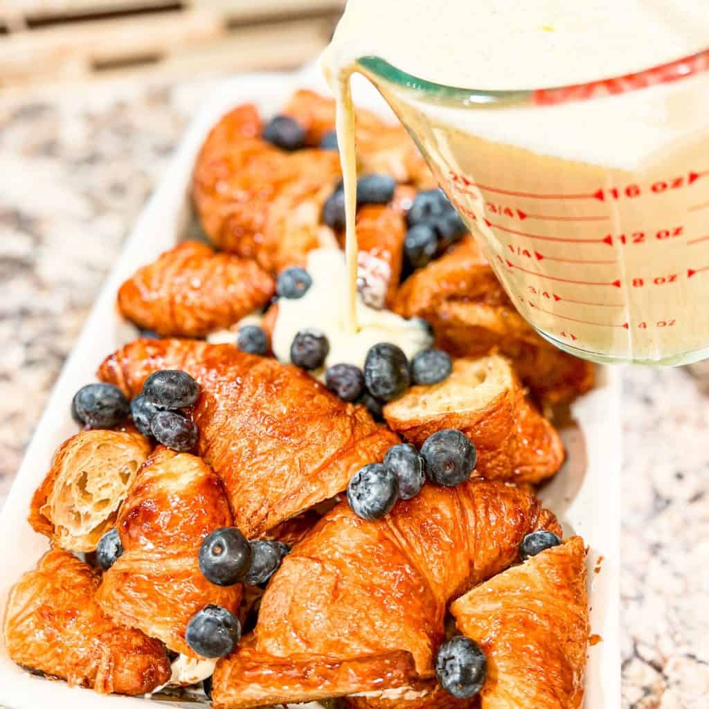 Pouring custard over croissants and blueberries in a white casserole dish.
