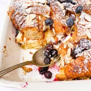 White casserole dish containing a croissant bread pudding with blueberries and almonds, with a spoon of berries.