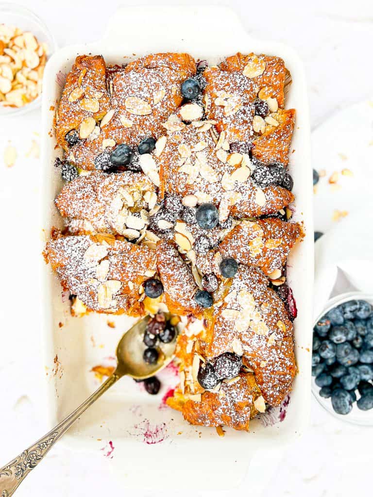 White casserole dish containing a croissant bread pudding with blueberries and almonds, with a spoon of berries.