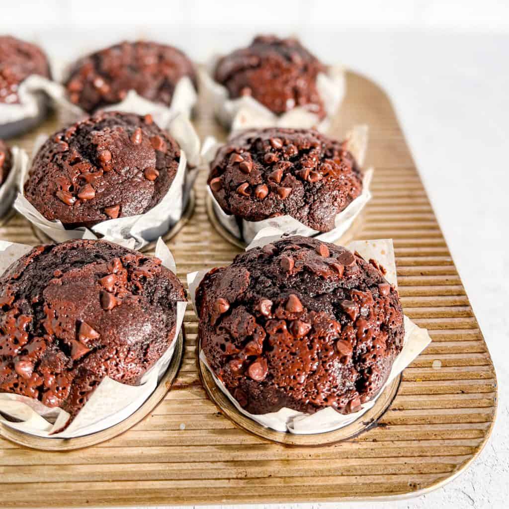 A muffin pan filled with Bakery Style Triple Chocolate Muffins in white parchment paper liners.
