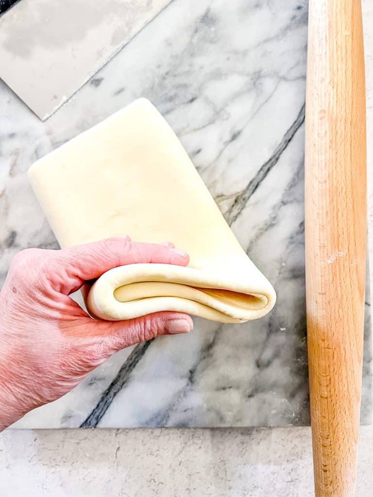 A hand holding a folded piece of Simple Rough Puff Pastry on a marble board with a rolling pin nearby.