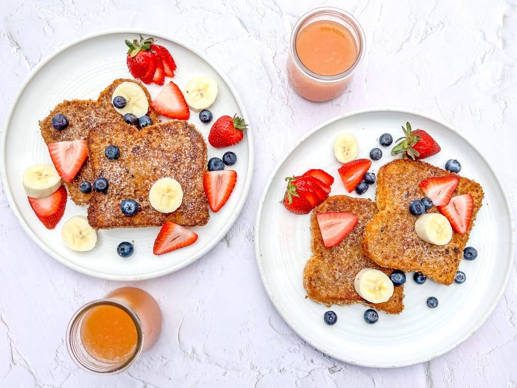 Two white plates of Cinnamon Toast Crunch French Toast with fruit and juice cups.