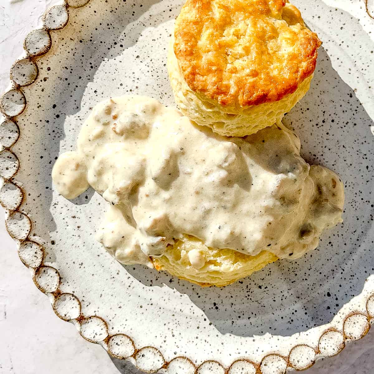 closeup of sausage gravy on a biscuit on a speckled white plate.