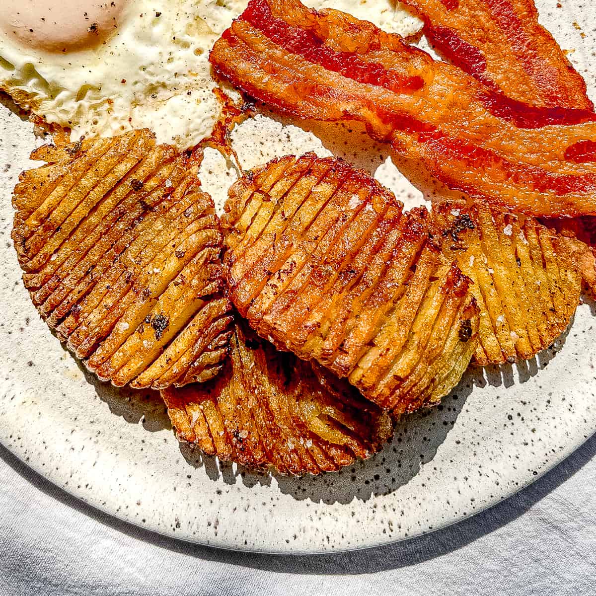 A white plate containing crispy accordion potatoes, bacon, and eggs.