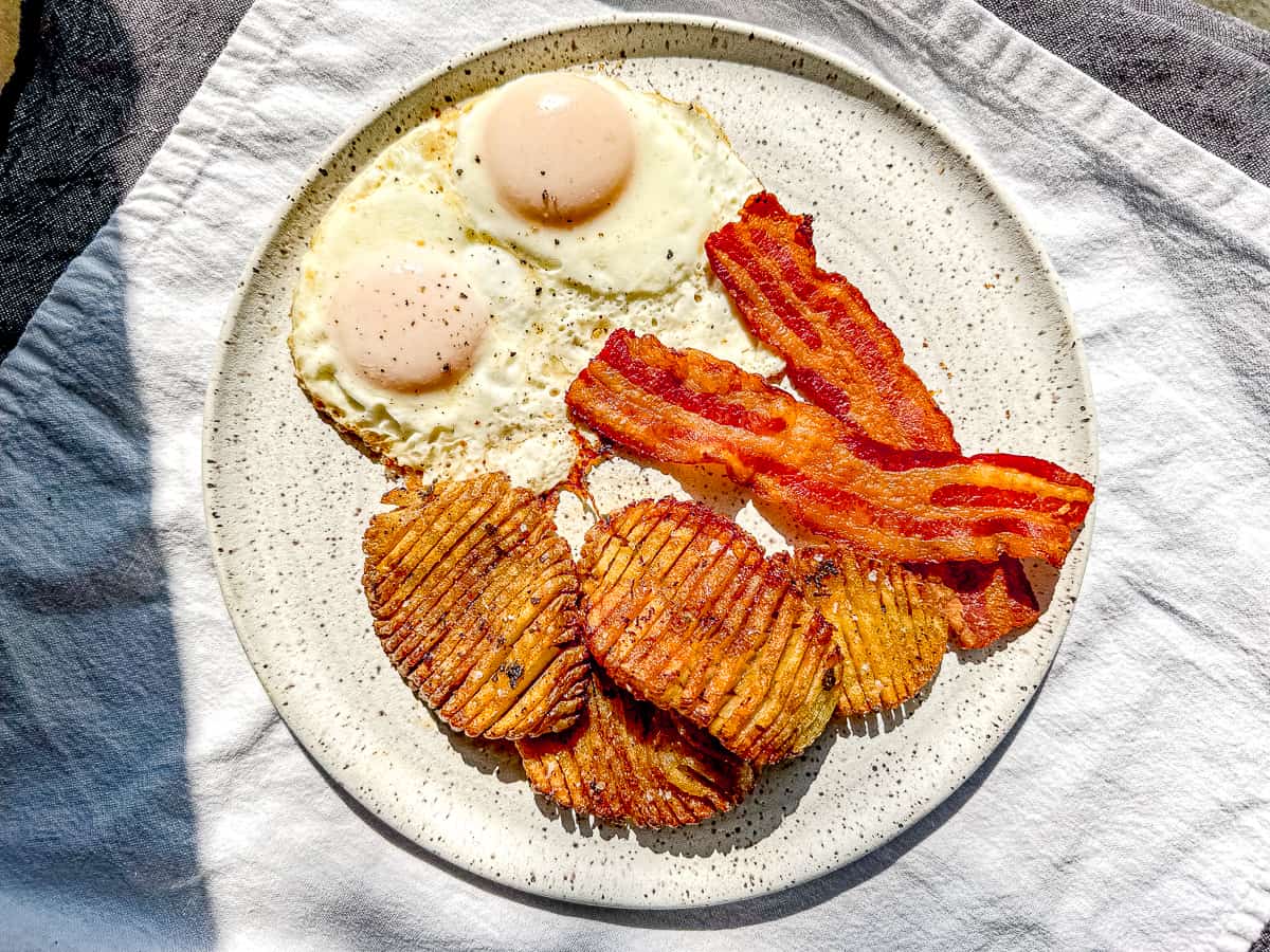 A white plate containing crispy accordion potatoes, bacon, and eggs.