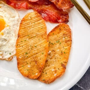 Two crispy accordion potatoes on a white plate with bacon and eggs.