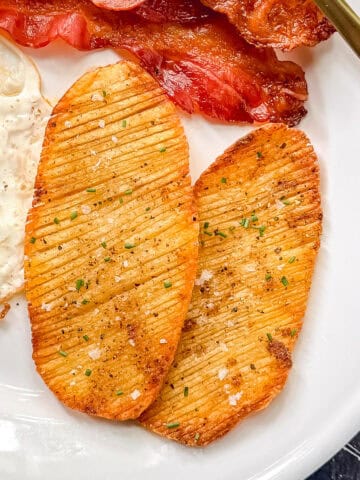 Two crispy accordion potatoes on a white plate with bacon and eggs.