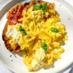 White plate containing a piece of toast with crispy prosciutto and creamy ricotta scrambled eggs.