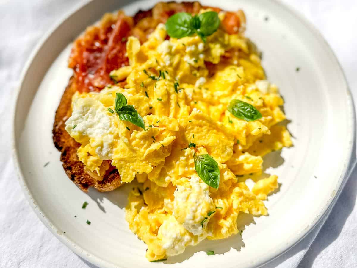 White plate containing a piece of toast with crsipy prosciutto and creamy ricotta scrambled eggs.