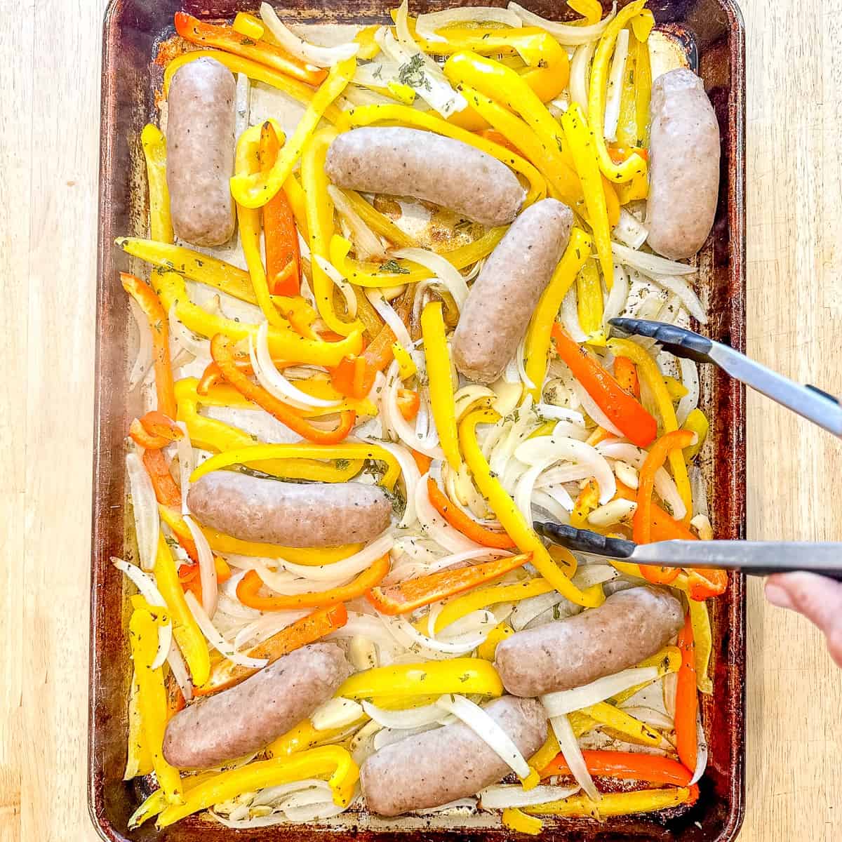 Sheet pan containing partially roasted sausage and peppers being turned.