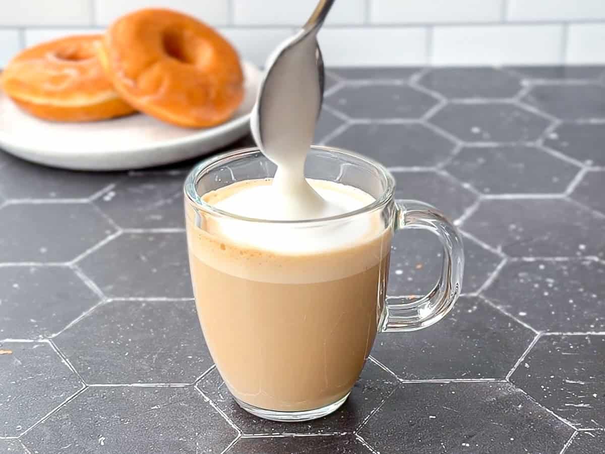 Adding foam to a donut latte with glazed donuts in the background.