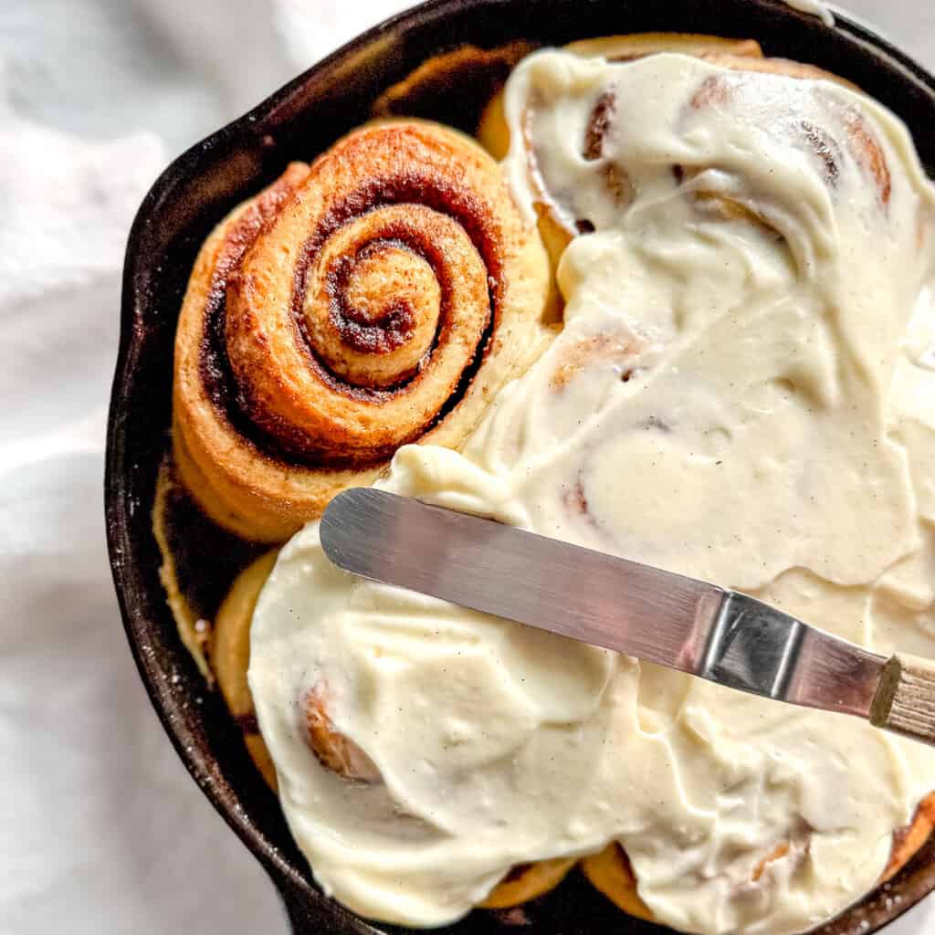 Spreading cream cheese frosting on a pan of cinnamon rolls.