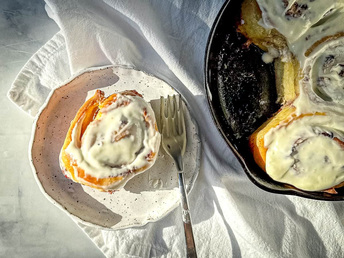A blueberry orange cinnamon roll on a white plate next to a cast-iron pan full of cinnamon rolls.