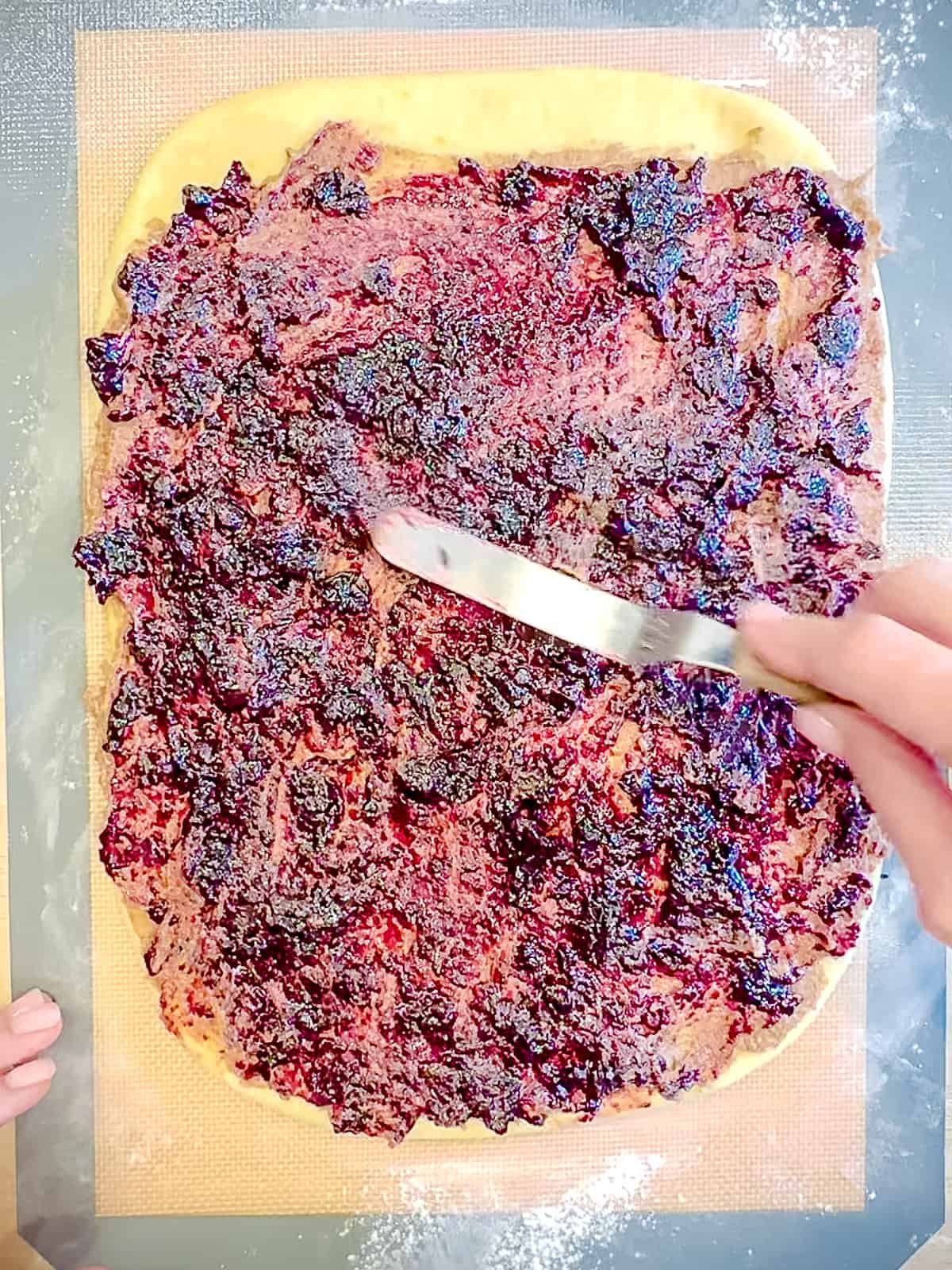 Spreading blueberry jam on top of cinnamon on blueberry cinnamon roll dough.