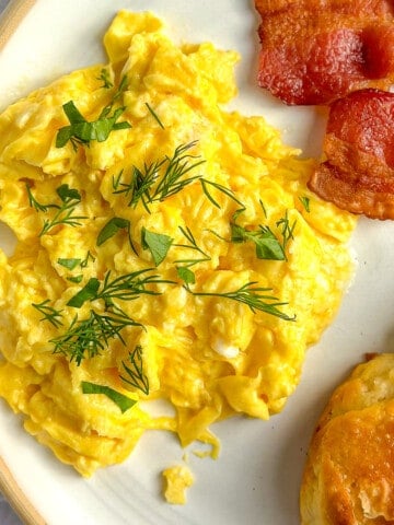Mascarpone scrambled eggs on a white plate with bacon and biscuits.