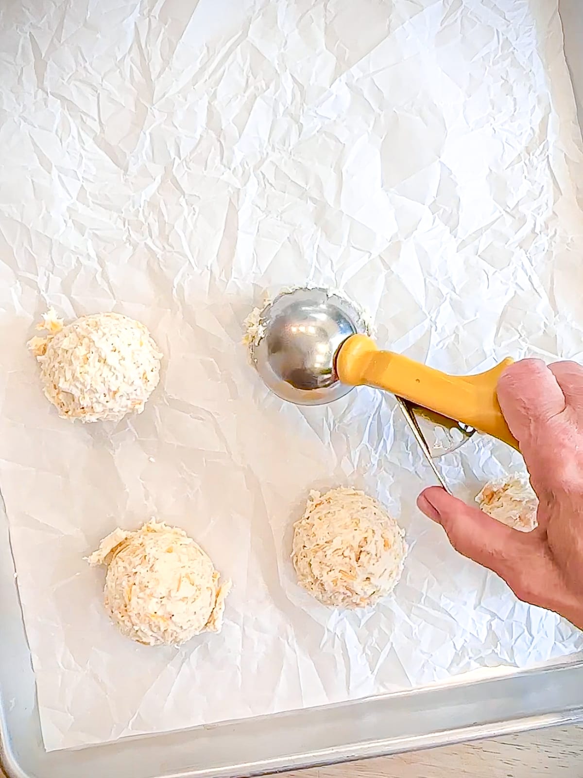 Dropping scoopfuls of biscuit dough onto parchment paper lined sheet pan.