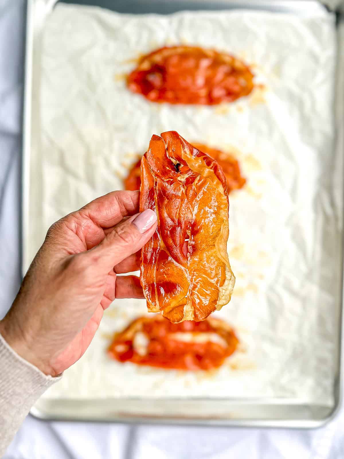A hand holding a piece of crispy prosciutto over a sheet tray of more.