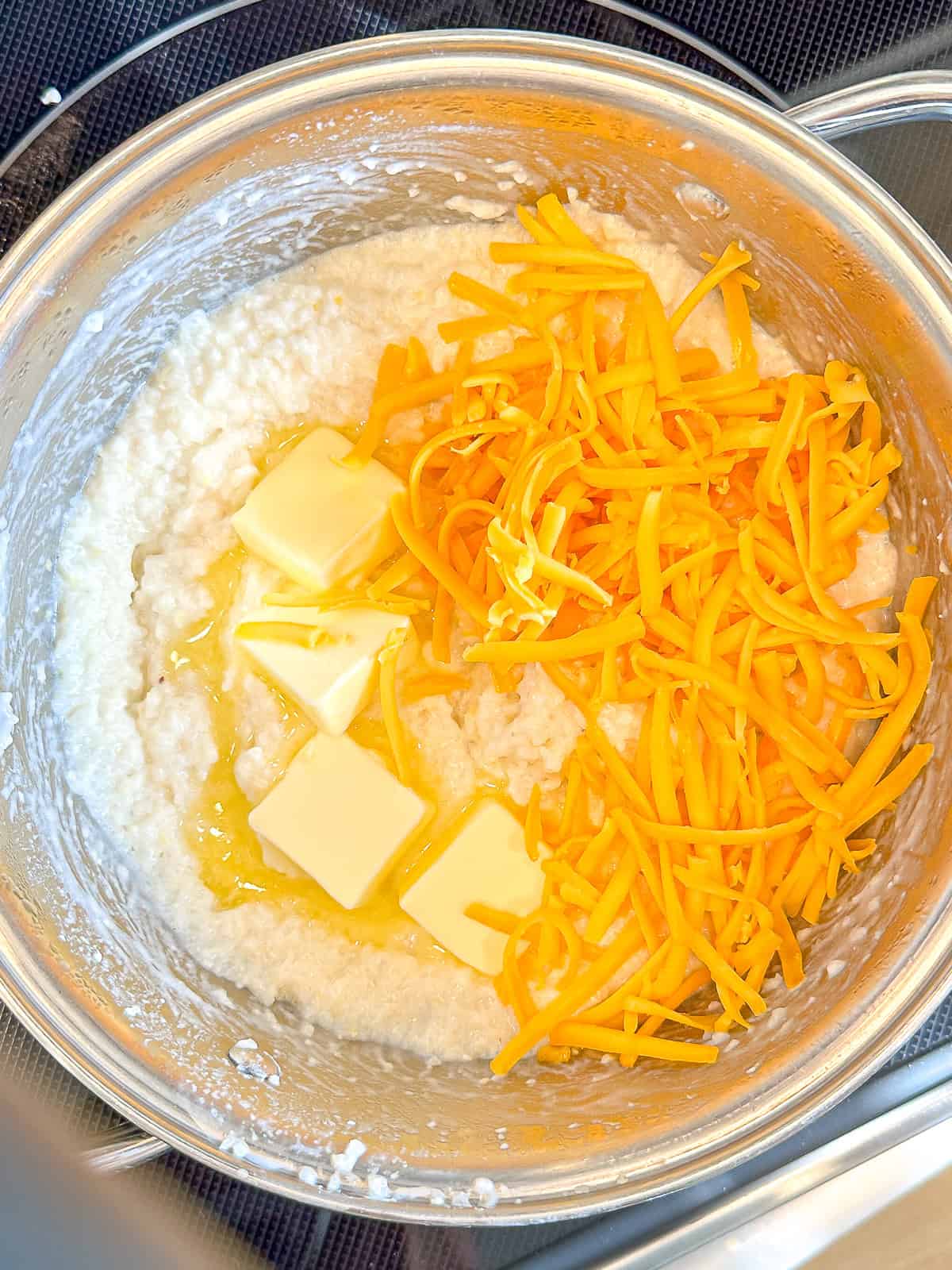 Adding pats of butter and shredded cheddar to grits cooking in a saucepan.