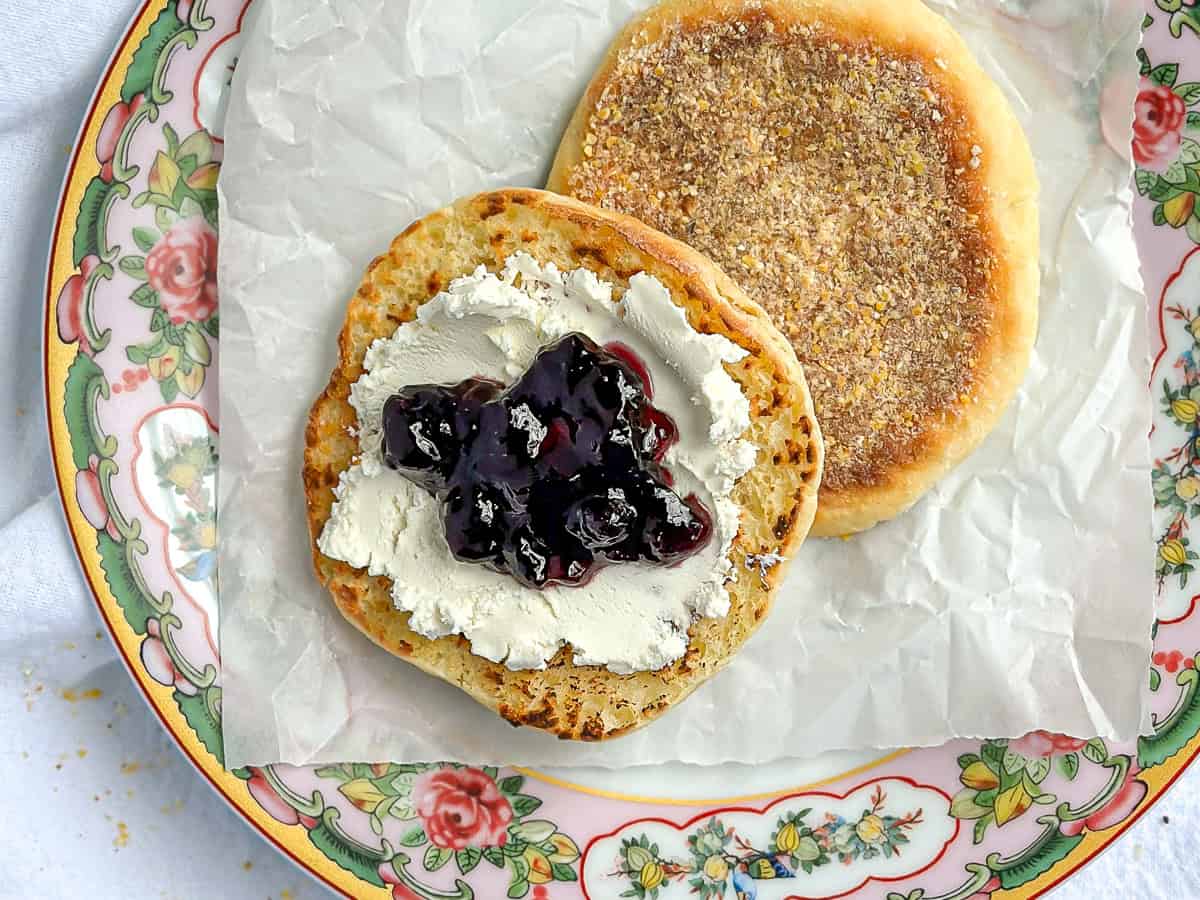 A toasted English muffin with cream cheese and blueberry jam on a floral plate.