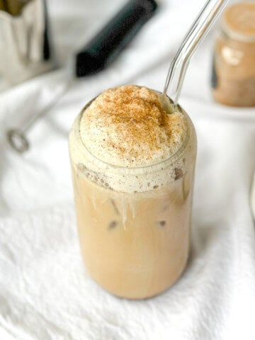 A glass of iced French toast latte with straw on a background of latte tools.