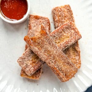 Cinnamon sugar French toast sticks on a cream plate with a dish of syrup.