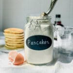 Homemade pancake mix in an airtight container next to a stack of pancakes.