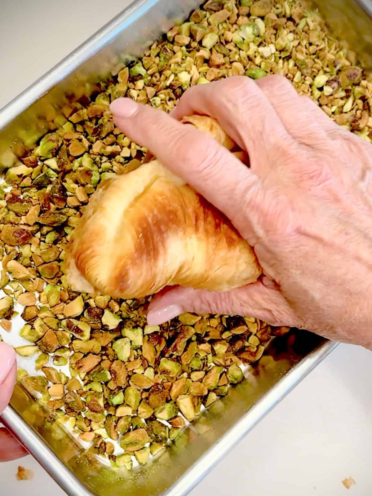 Pressing the tops of unbaked pistachio croissants in a tray of chopped pistachios.